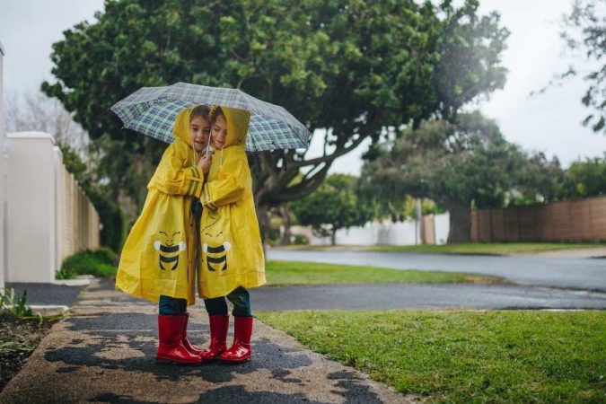 Deux jeunes enfants, complices et souriants, partagent le même parapluie et portent des imperméables jaunes ainsi que des bottes rouges, défiant joyeusement le mauvais temps pour s'occuper les enfants les jours de pluie.